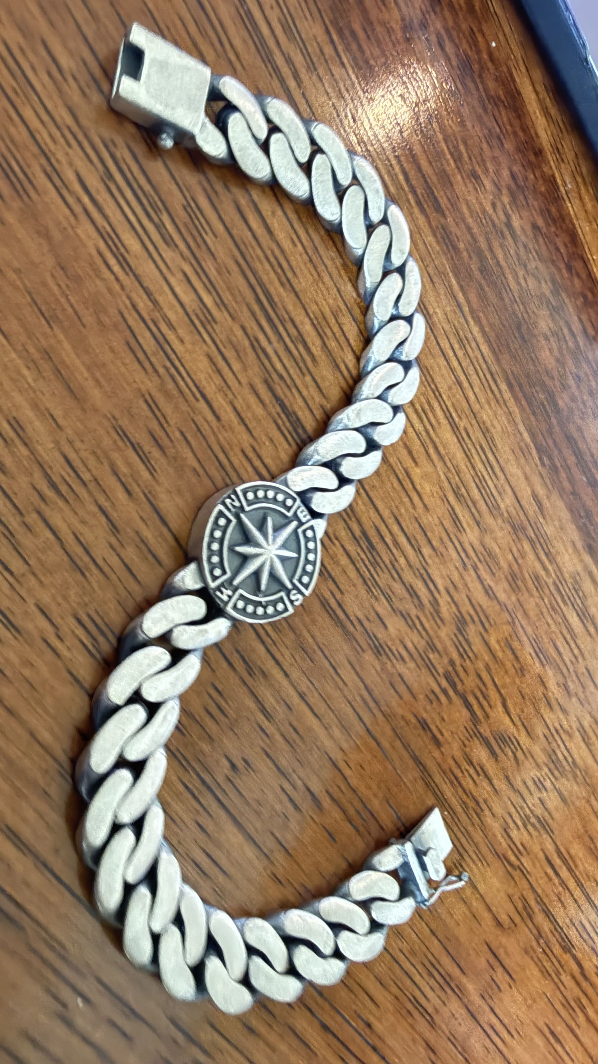 A silver-toned Cuban link chain bracelet with a circular medallion charm featuring a compass design, displayed on a wooden surface.