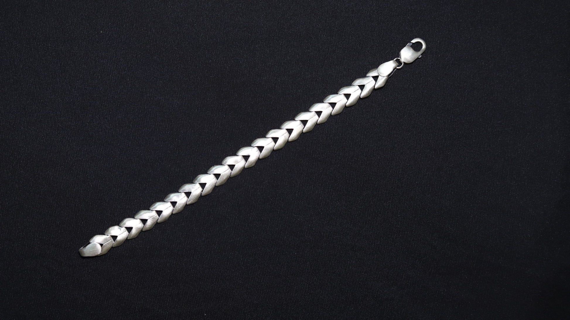 A silver-toned braided chain bracelet with a lobster clasp, displayed diagonally on a solid black background.