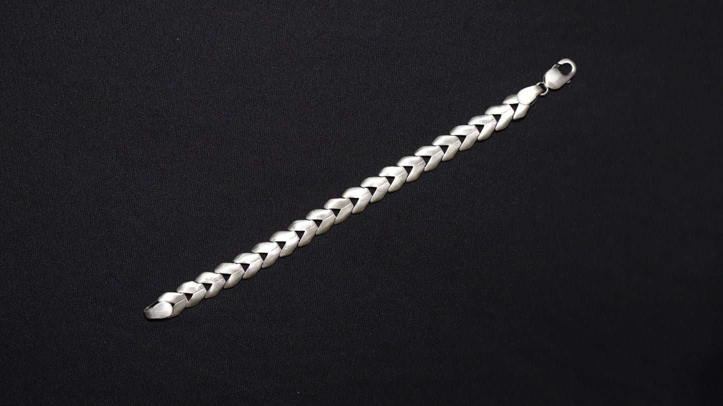 A silver braided chain bracelet with a lobster clasp, displayed diagonally on a solid black background.