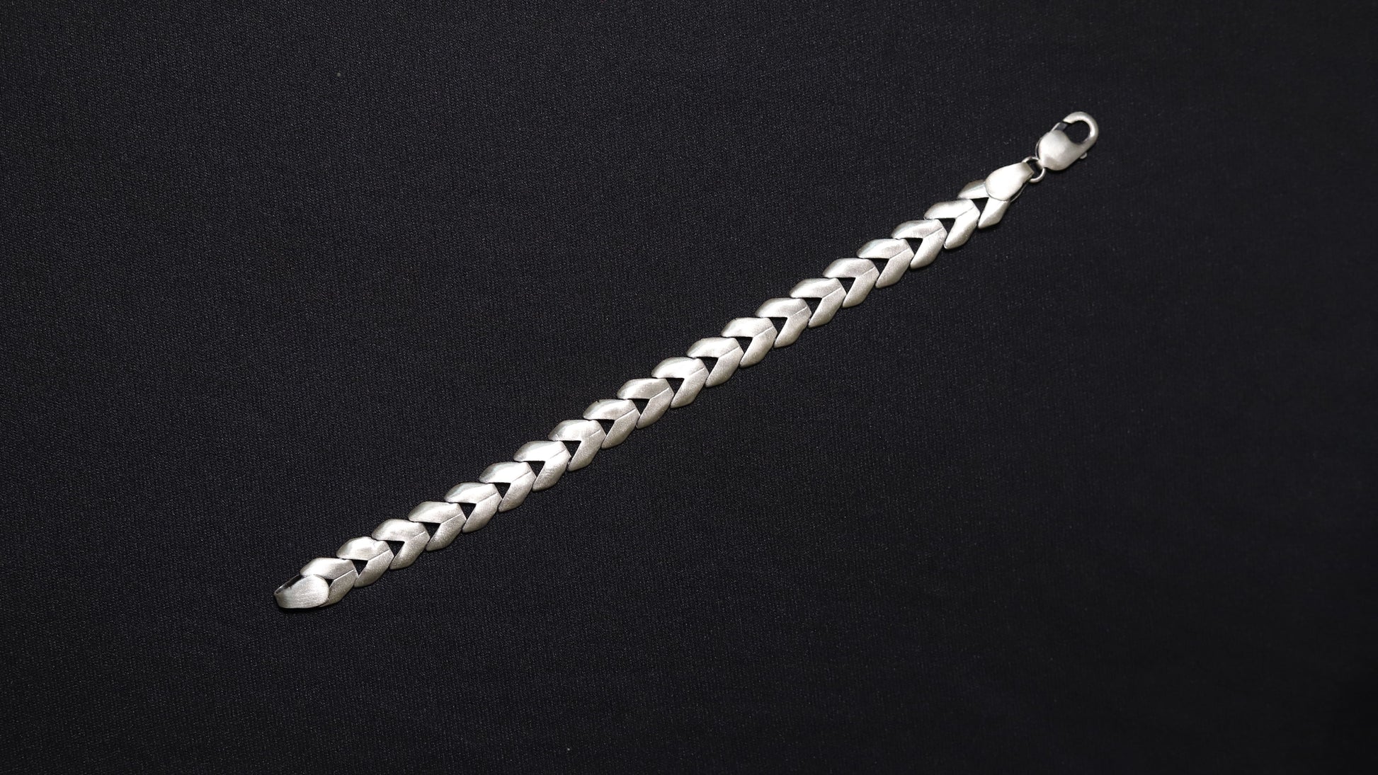 A silver braided chain bracelet with a lobster clasp, displayed diagonally on a solid black background.