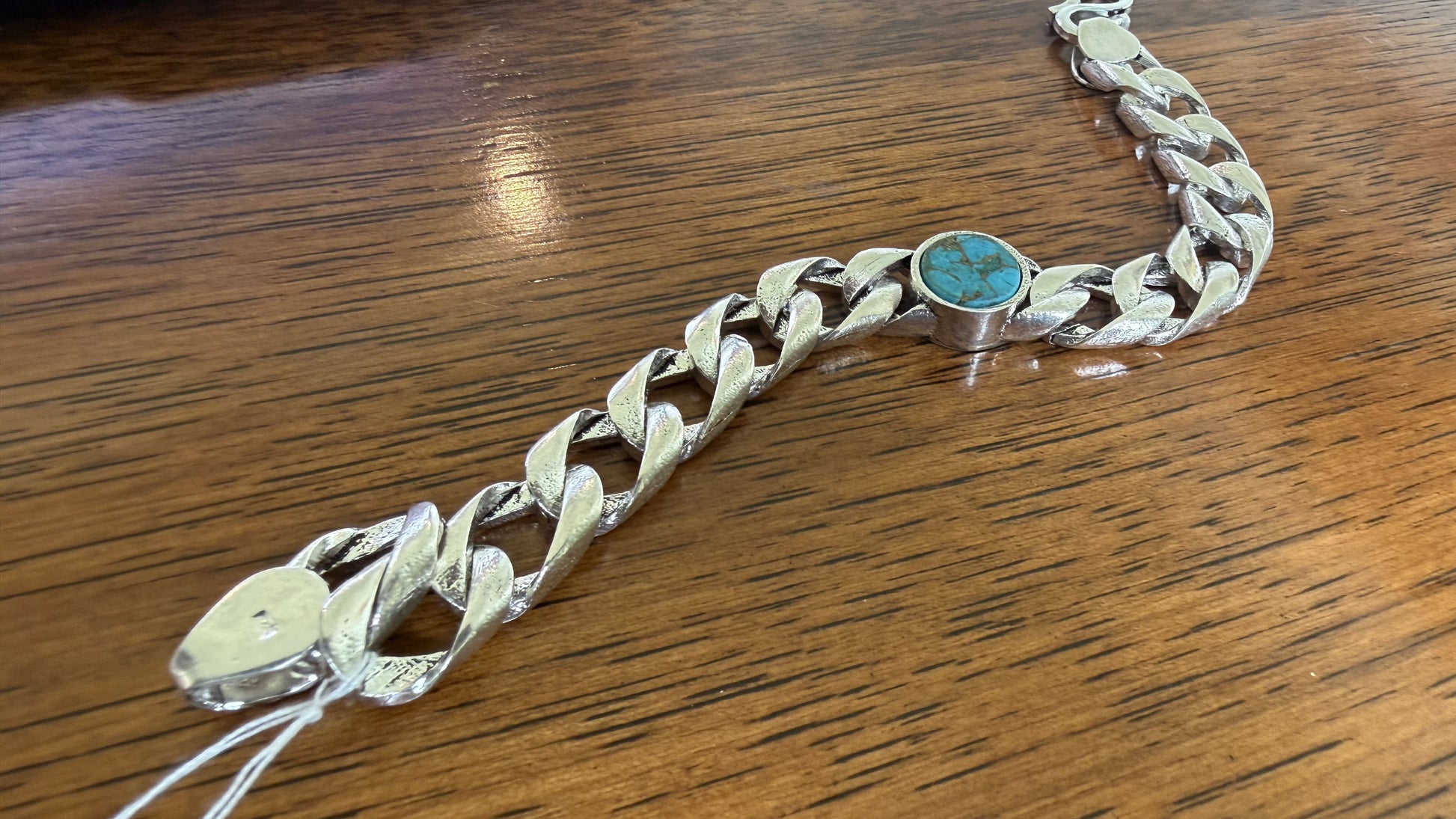 A silver-toned curb link bracelet featuring a round turquoise stone centerpiece, displayed on a wooden surface.