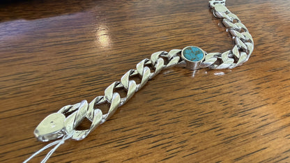 A silver-toned curb link bracelet featuring a round turquoise stone centerpiece, displayed on a wooden surface.