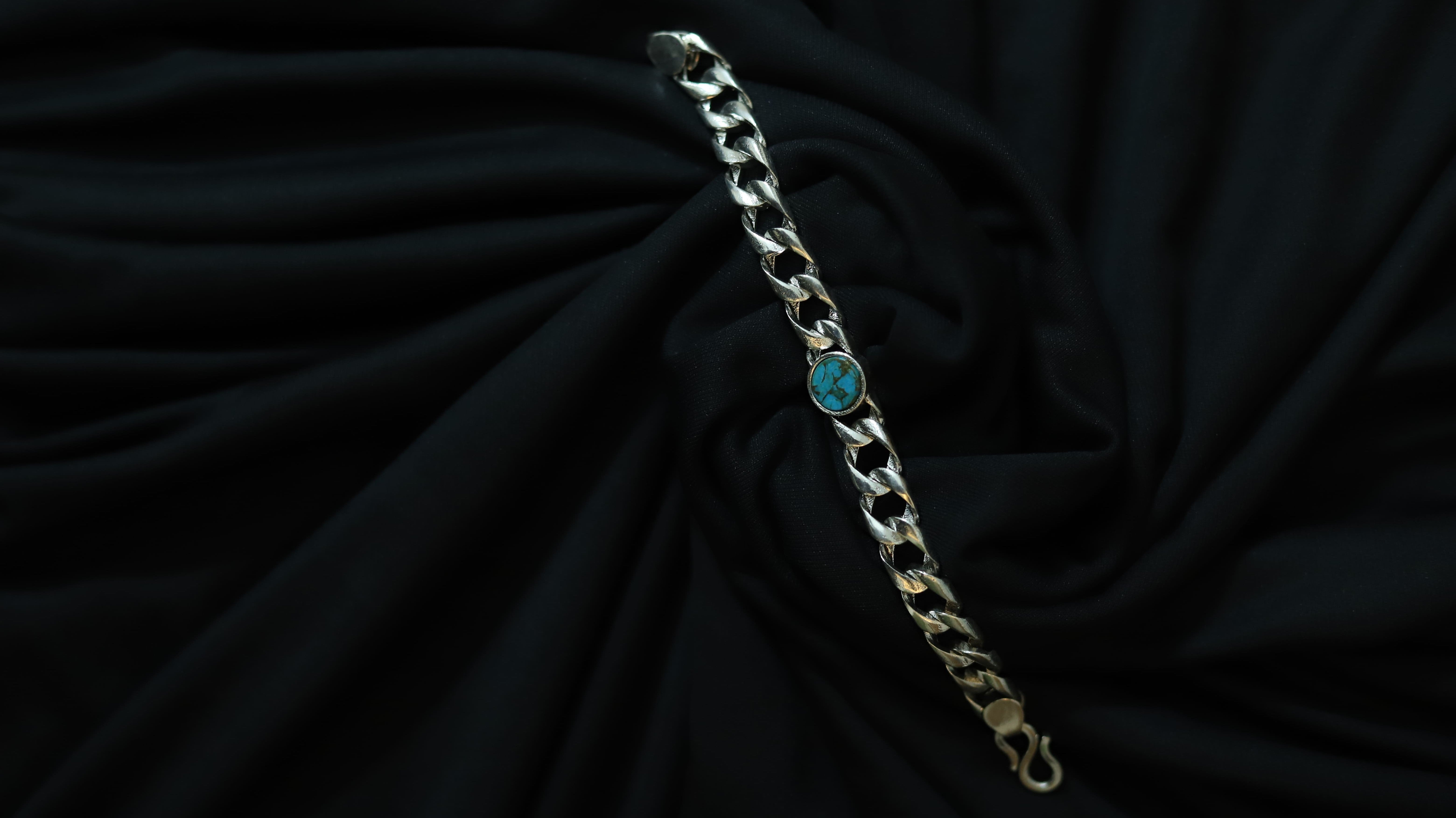 A silver-toned curb chain bracelet with a small round turquoise blue stone accent, displayed on a draped black fabric background.