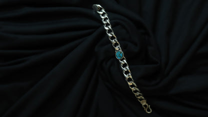 A silver-toned curb chain bracelet with a small round turquoise blue stone accent, displayed on a draped black fabric background.