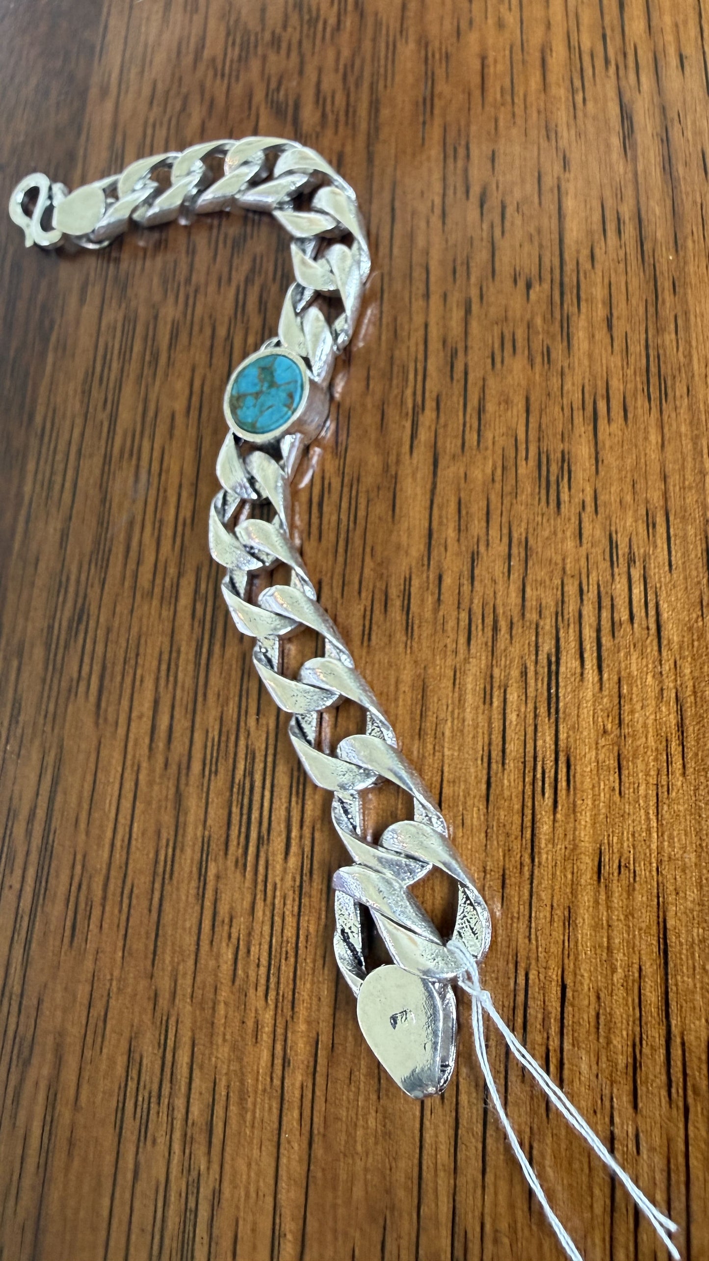 A silver-toned Cuban link chain bracelet with a round turquoise stone inlay, displayed on a brown wooden surface.
