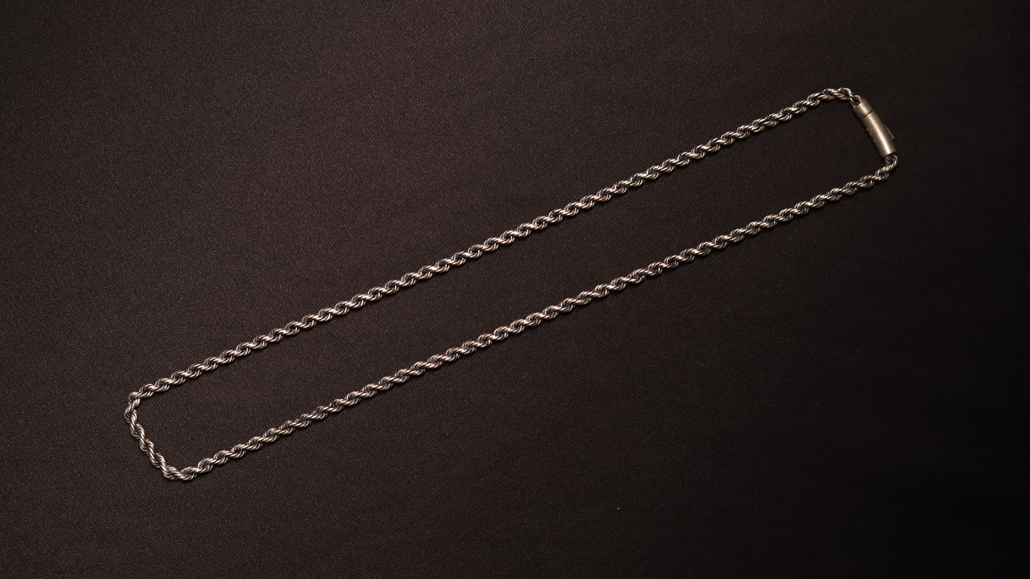 A silver-toned twisted rope chain necklace with a simple rectangular clasp, laid flat on a dark textured background.