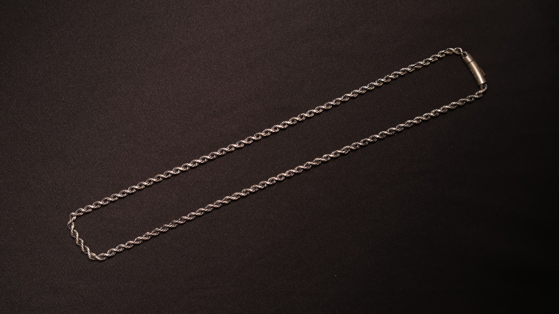 A silver-toned twisted rope chain necklace with a simple rectangular clasp, laid flat on a dark textured background.