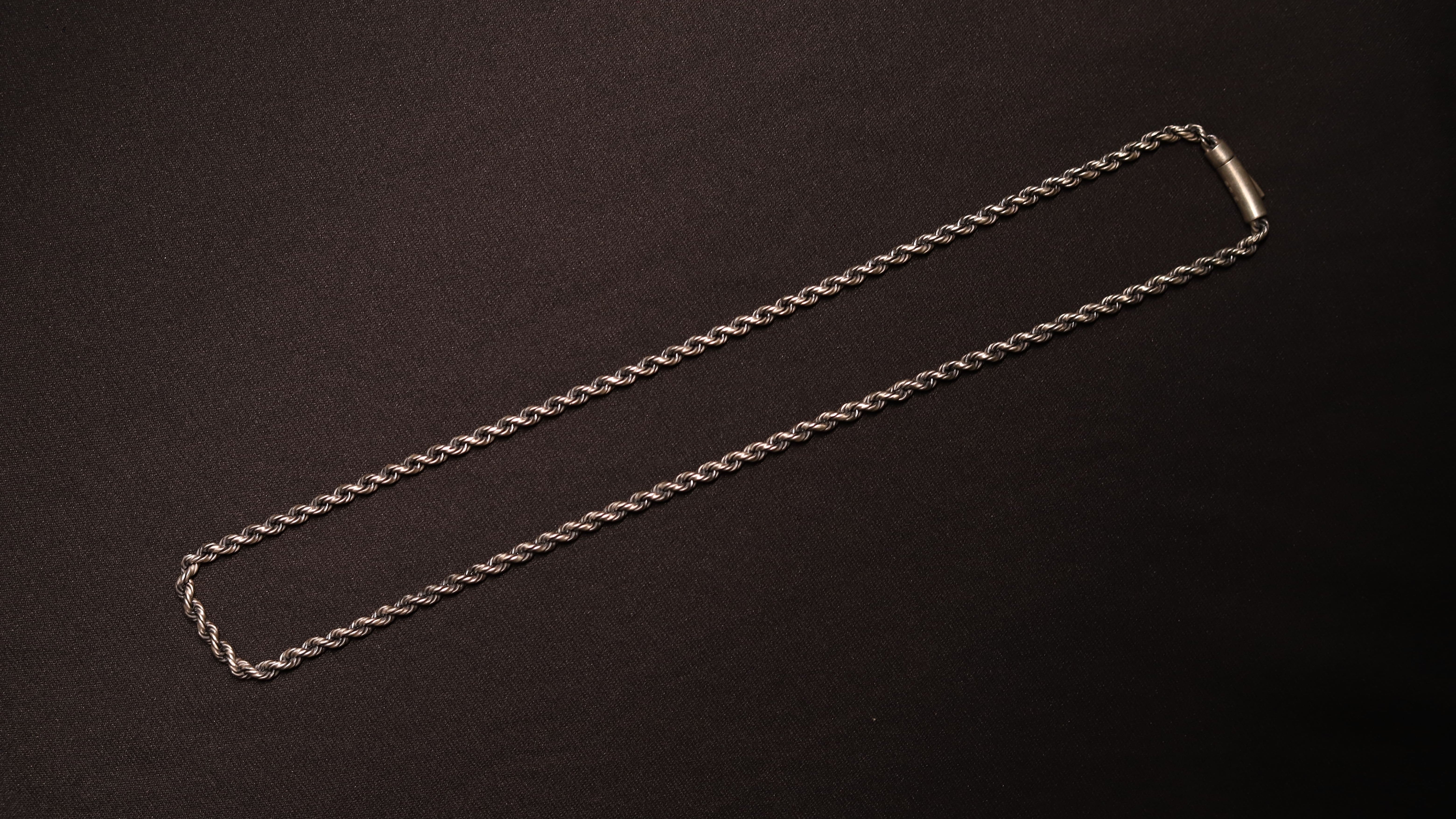 A silver-toned twisted rope chain necklace with a simple rectangular clasp, laid flat on a dark textured background.