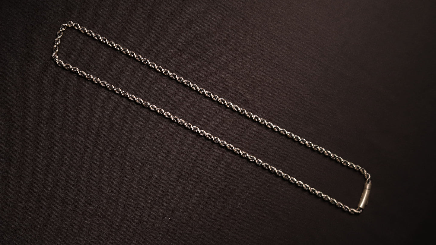 A silver twisted rope chain necklace with a polished rectangular clasp, laid out diagonally on a dark brown textured background.
