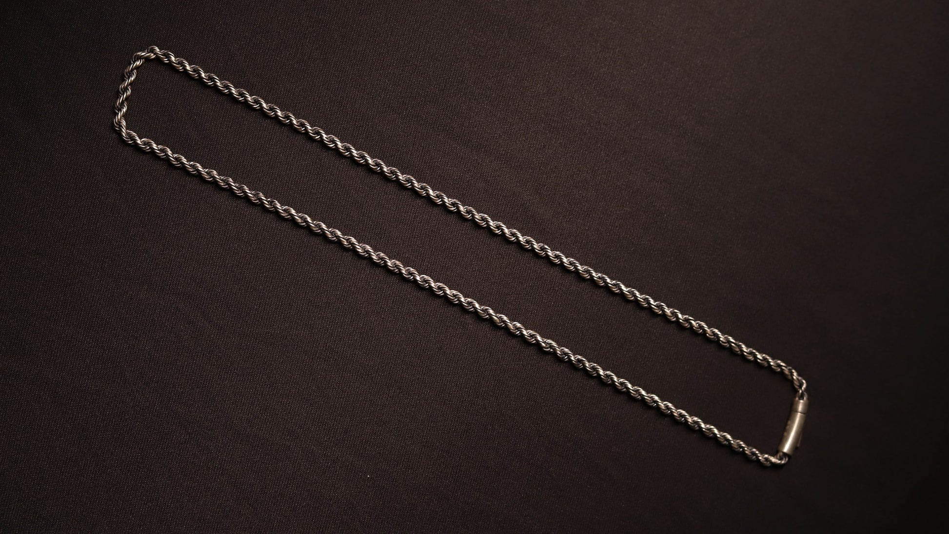 A silver twisted rope chain necklace with a polished rectangular clasp, laid out diagonally on a dark brown textured background.