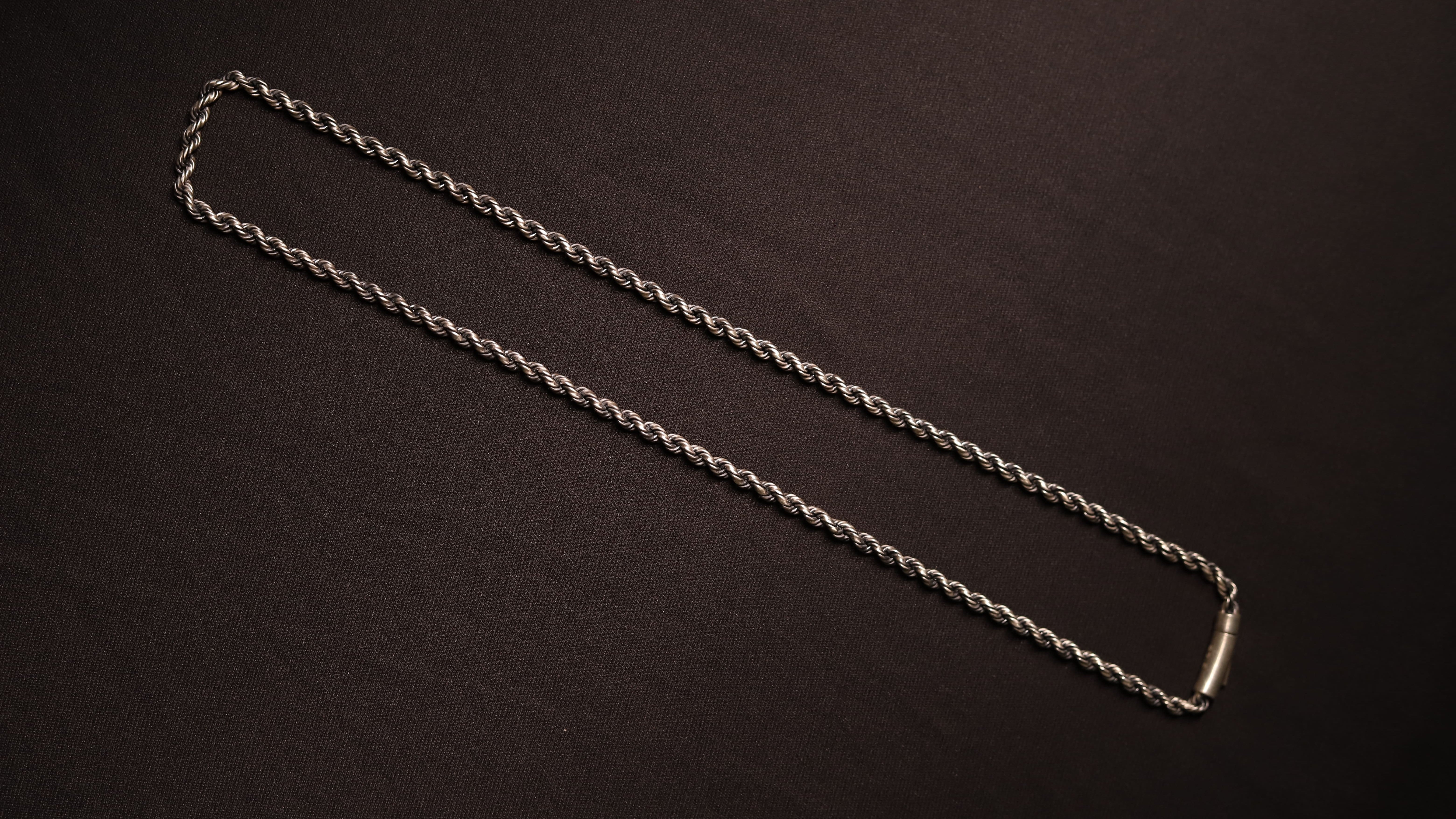 A silver twisted rope chain necklace with a polished rectangular clasp, laid out diagonally on a dark brown textured background.