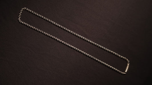 A silver twisted rope chain necklace with a polished rectangular clasp, laid out diagonally on a dark brown textured background.