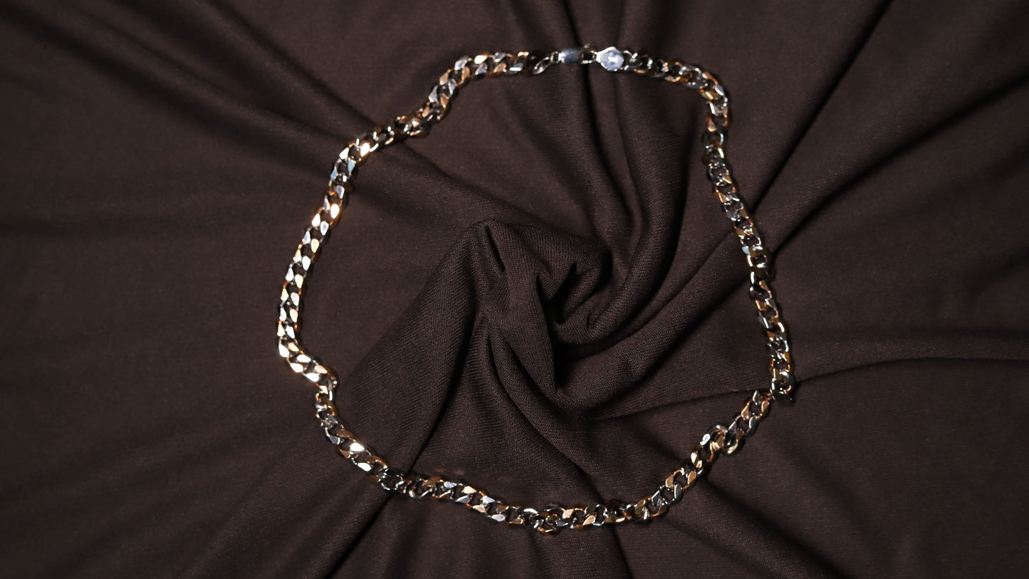 A chunky silver-toned curb chain necklace arranged in a circle on a dark brown draped fabric background.