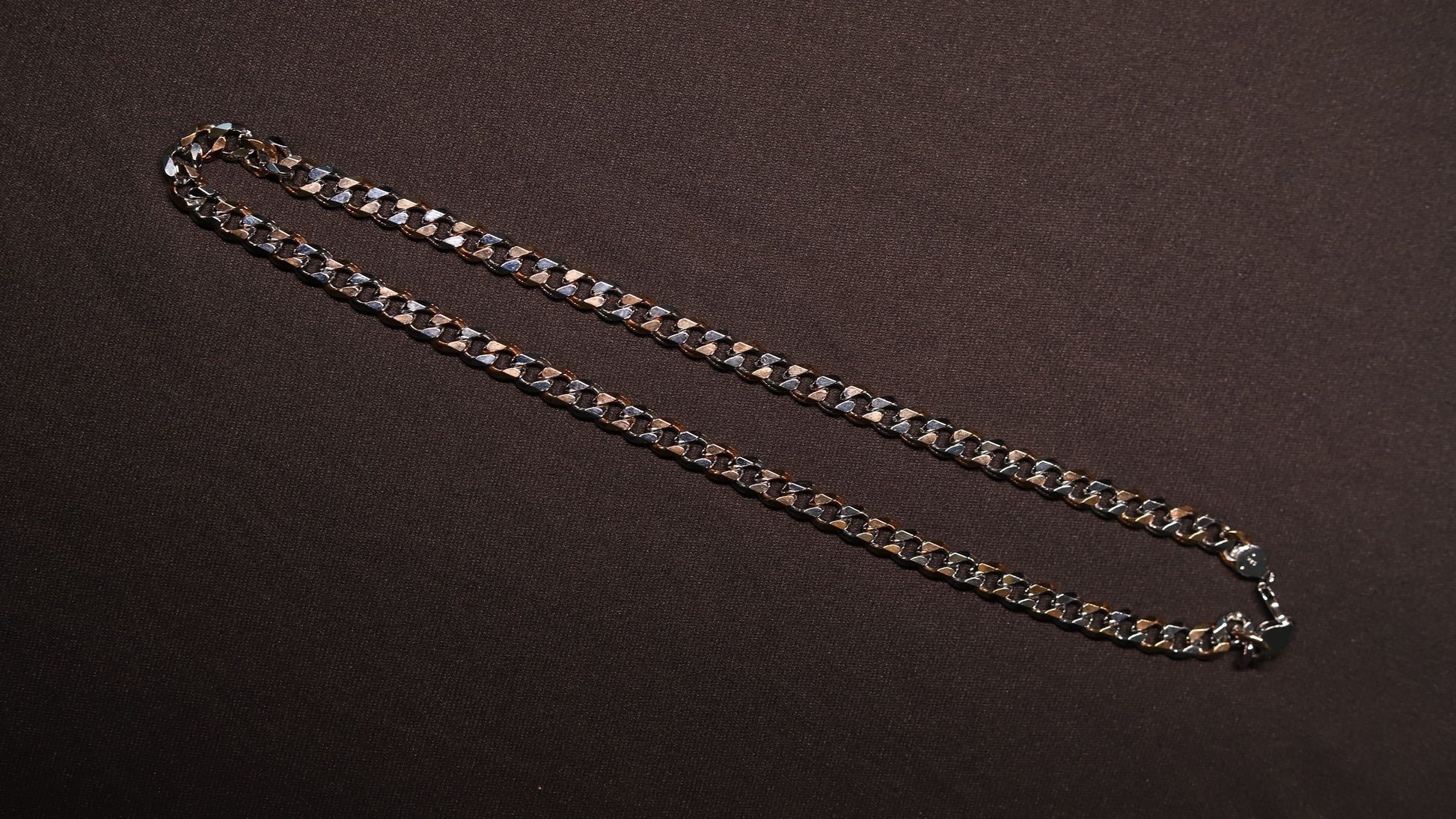 A silver-toned metal curb chain necklace laid out diagonally on a dark brown textured background.