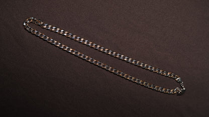 A silver-toned metal curb chain necklace laid out diagonally on a dark brown textured background.
