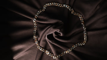 A chunky gold-toned metal chain necklace arranged in a circle on dark brown draped fabric.