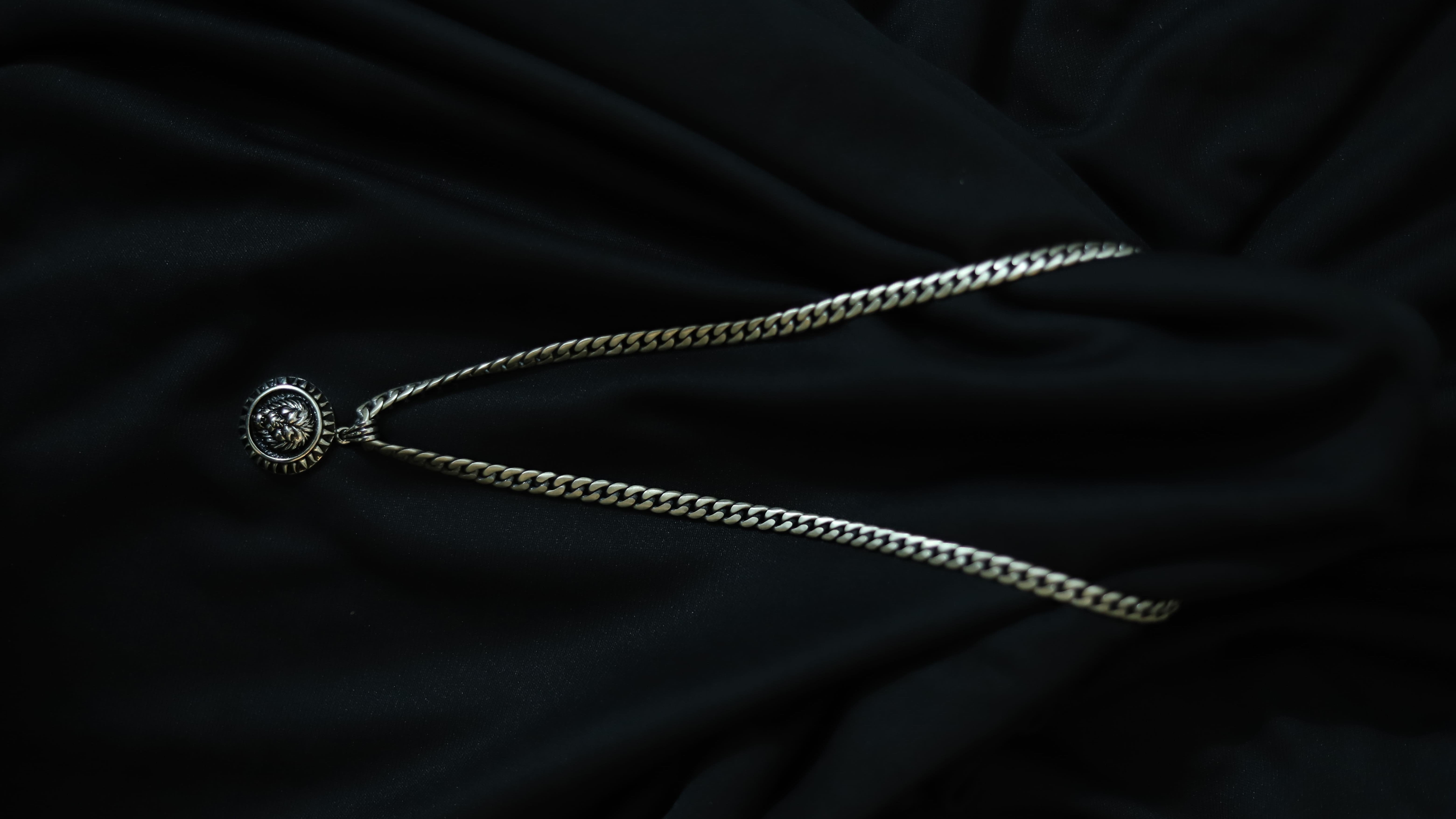 A silver-toned chain necklace with a round medallion pendant featuring a stylized 'V' emblem, displayed on a draped black fabric background.