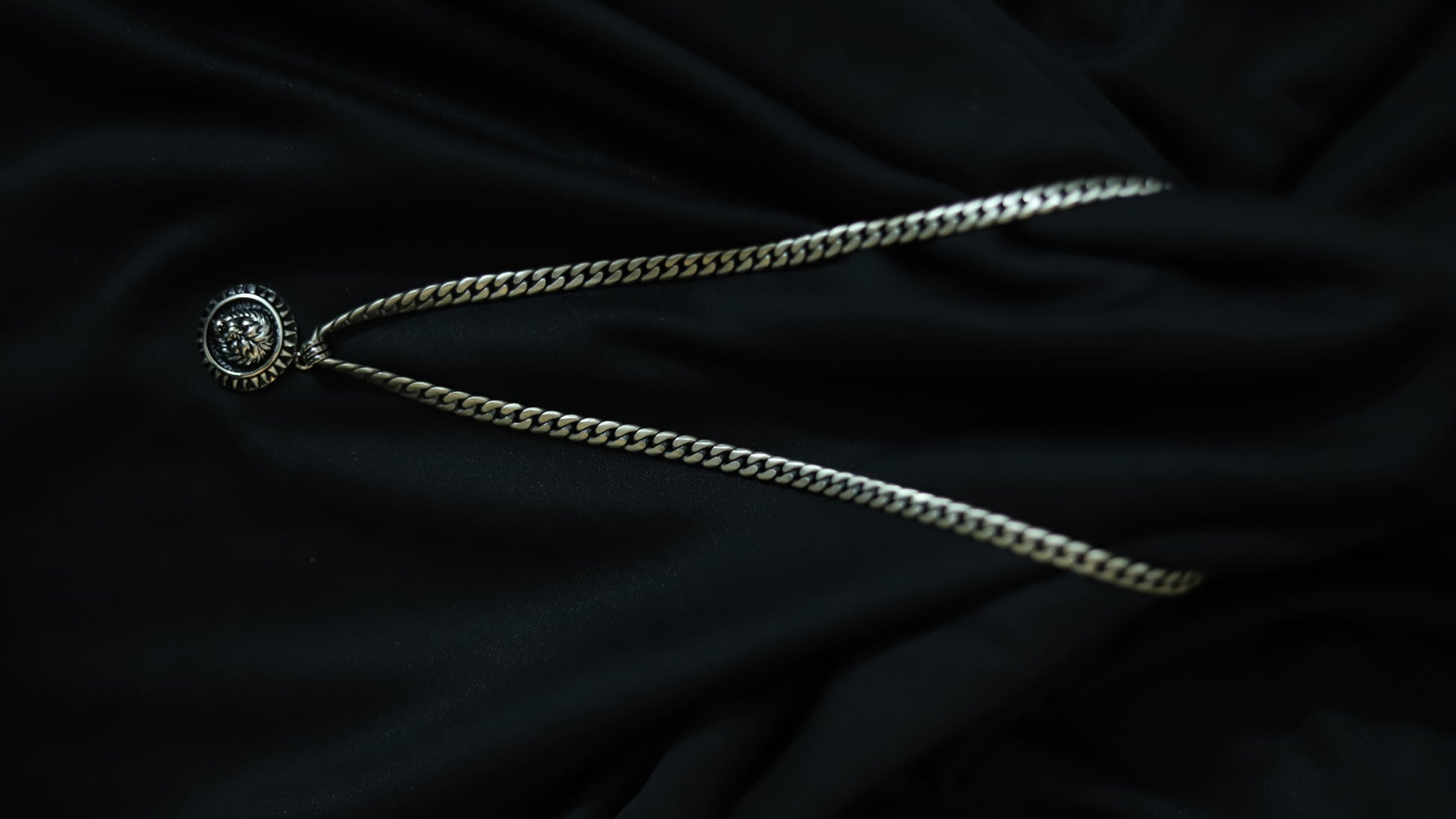 A silver-toned chain necklace with a circular medallion pendant featuring a stylized face, laid out on a dark black fabric background.