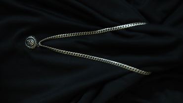 A silver-toned chain necklace with a circular medallion pendant featuring a stylized face, laid out on a dark black fabric background.