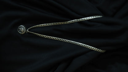 A silver-toned chain necklace with a circular medallion pendant featuring a stylized face, laid out on a dark black fabric background.