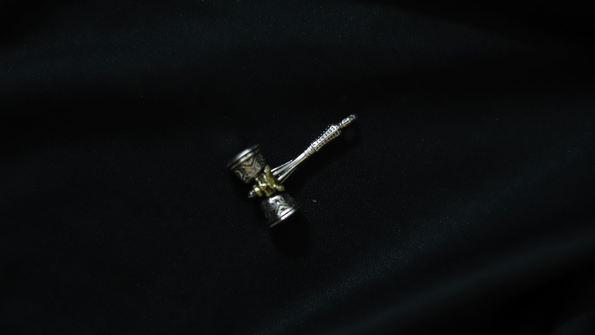 A miniature hammer-shaped accessory with a silver-toned handle and head, featuring gold-toned detailing, resting on a dark black fabric background.