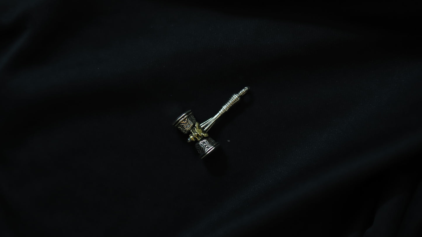 A small silver-toned metal pendant or charm with a textured cylindrical head and a long pointed stem, photographed on a dark black fabric background.