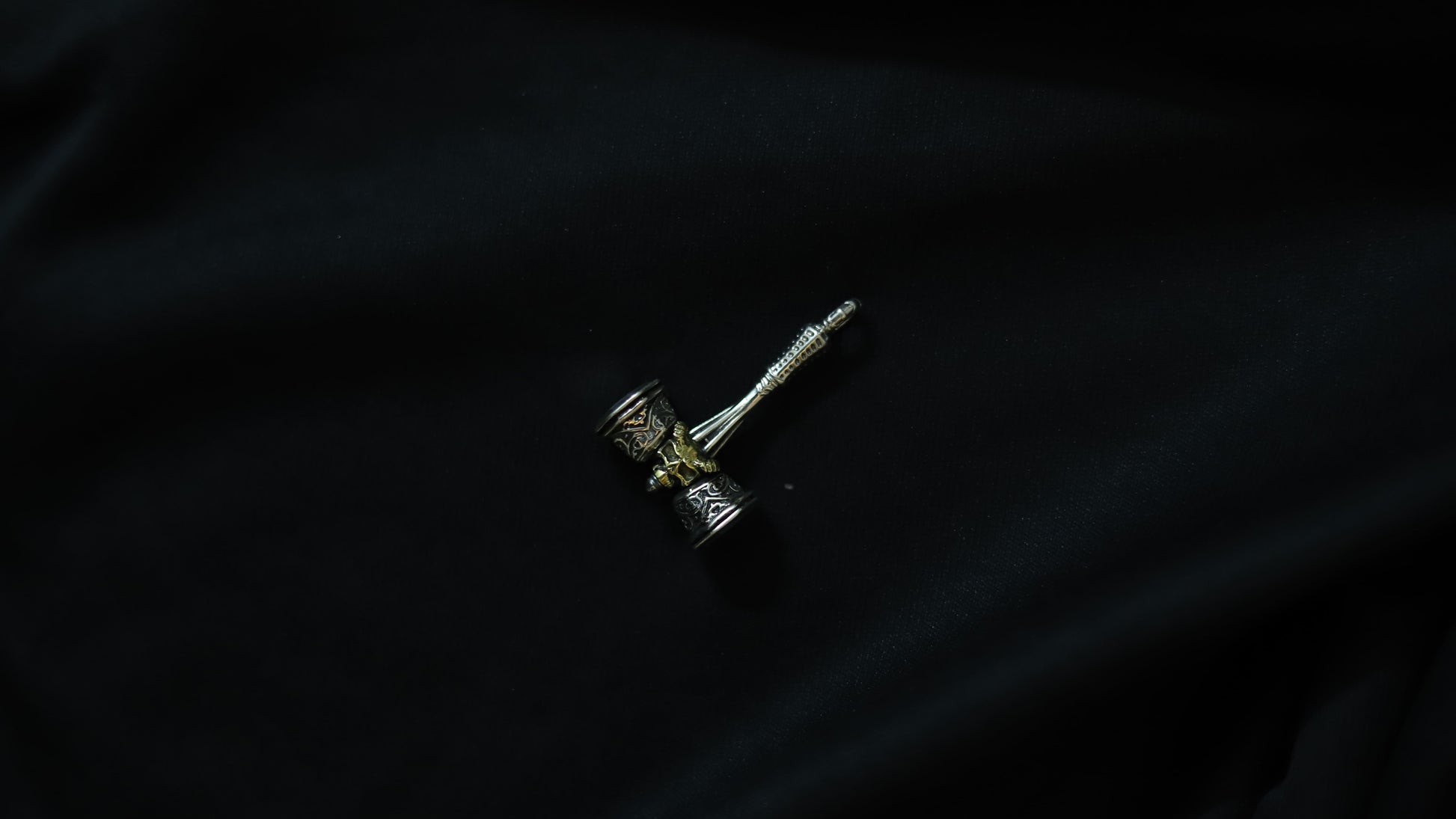 A small silver-toned metal pendant or charm with a textured cylindrical head and a long pointed stem, photographed on a dark black fabric background.