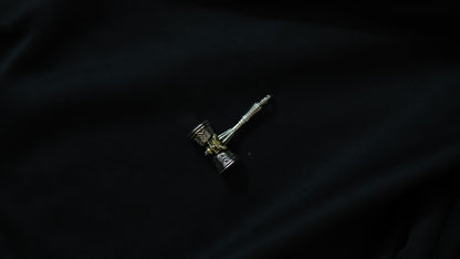 A small silver-toned metal pendant or charm with a textured cylindrical head and a long pointed stem, photographed on a dark black fabric background.