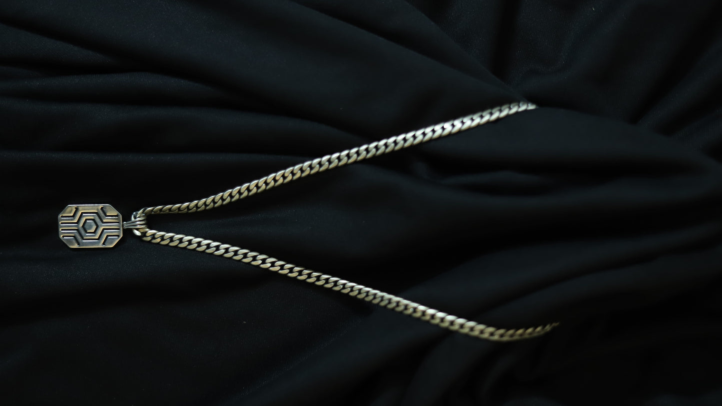 A silver-toned chain necklace with a geometric patterned pendant, laid out on a draped black fabric background.