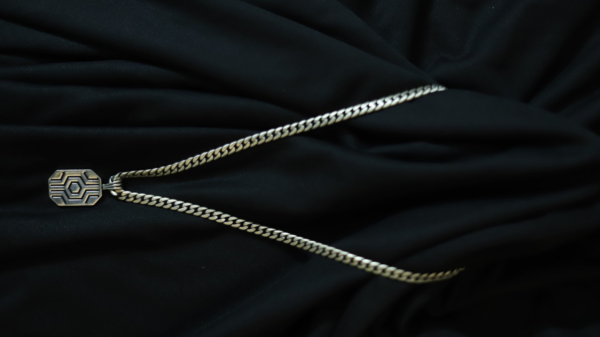 A silver-toned chain necklace with a geometric patterned pendant, laid out on a draped black fabric background.