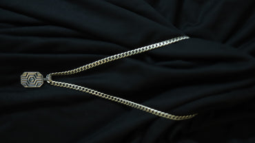 A silver-toned chain necklace with a geometric patterned pendant, laid out on a draped black fabric background.
