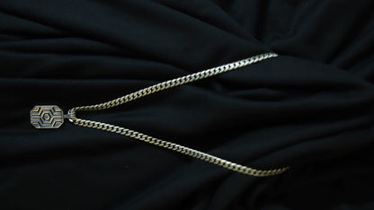 A silver-toned chain necklace with a geometric patterned pendant, laid out on a draped black fabric background.