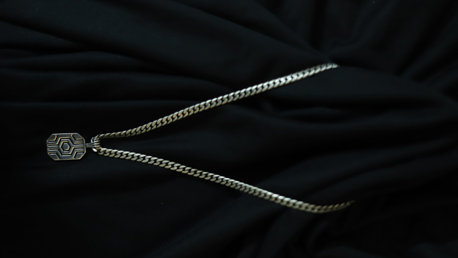 A silver-toned chain necklace with a geometric, hexagonal pendant featuring a sunburst pattern, displayed on a draped black fabric background.