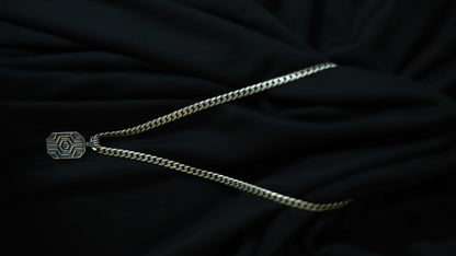 A silver-toned chain necklace with a geometric, hexagonal pendant featuring a sunburst pattern, displayed on a draped black fabric background.