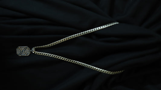 A silver-toned chain necklace with a geometric, hexagonal pendant featuring a sunburst pattern, displayed on a draped black fabric background.