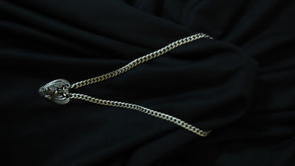 A silver-toned chain necklace with a textured, decorative oval pendant, laid out on a draped black fabric background.