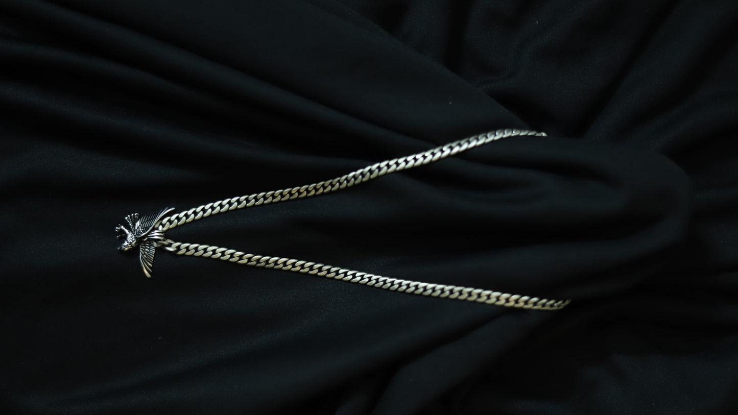 A silver-toned metal chain necklace with a small winged bird pendant, laid out on a draped black fabric background.