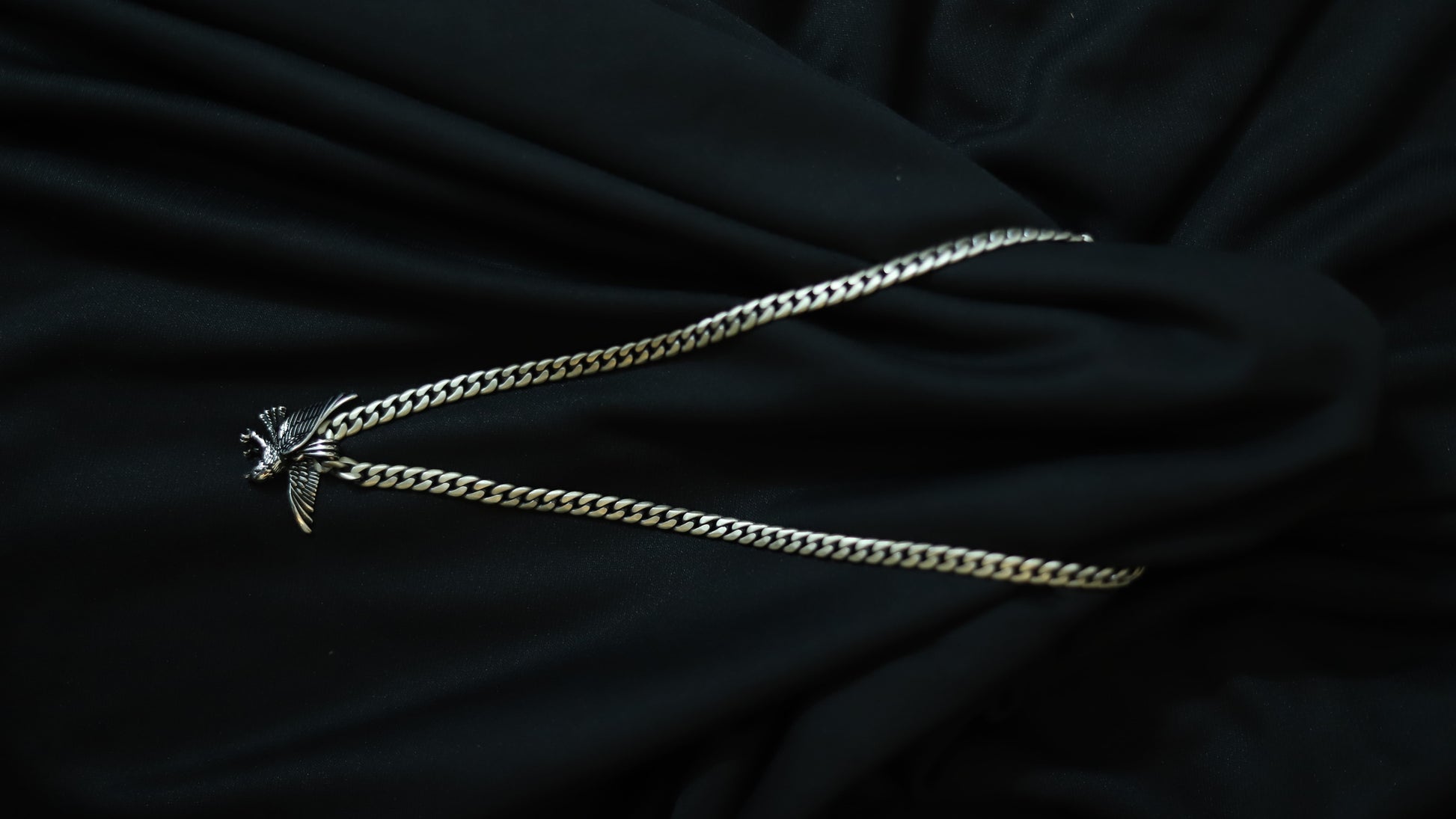 A silver-toned curb chain necklace with a small bird pendant, laid out on draped black fabric.