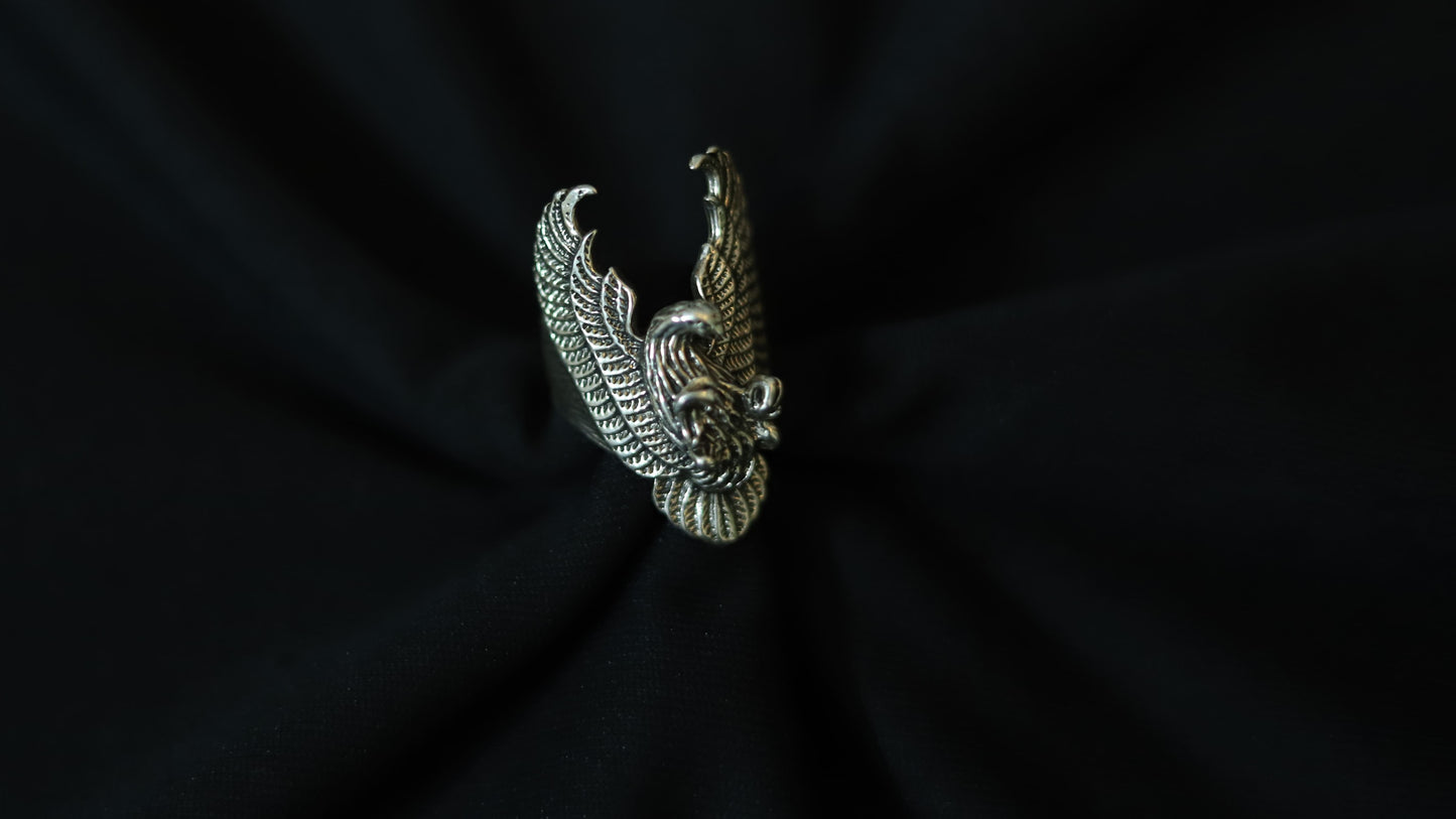 A silver-toned metal ring featuring a detailed, textured design of a bird with spread wings, displayed on a draped black fabric background.