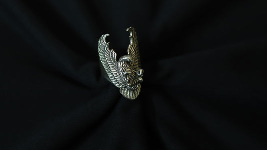 A silver-toned metal ring featuring a detailed, textured design of a bird with spread wings, displayed on a draped black fabric background.