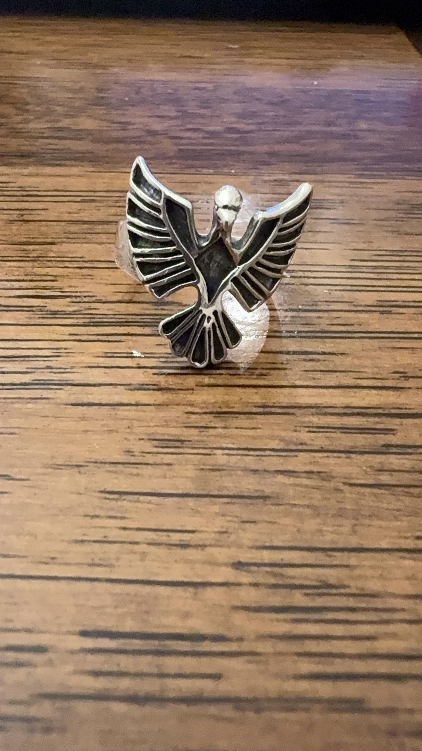 A silver-toned metal ring shaped like a stylized bird or winged emblem, photographed on a wooden tabletop.