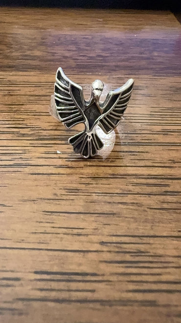 A silver-toned metal ring shaped like a stylized bird or winged emblem, photographed on a wooden tabletop.