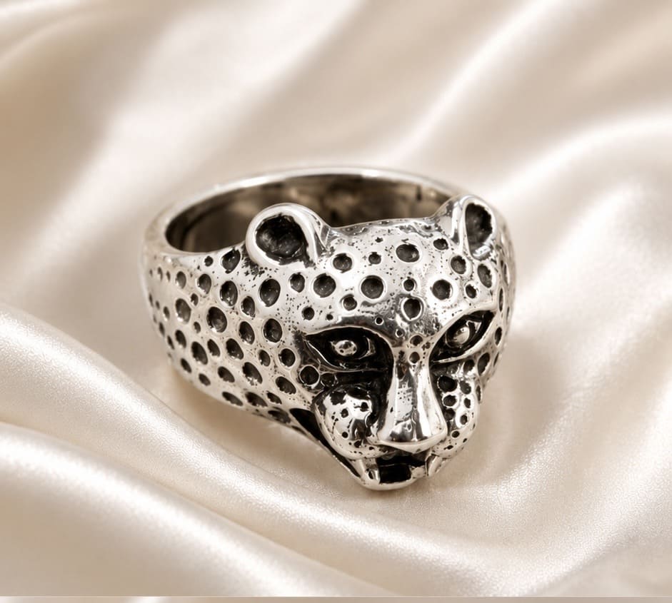A sterling silver ring shaped like a leopard head, featuring a textured spotted pattern and small clear stones in the eyes, displayed on a draped cream satin fabric.