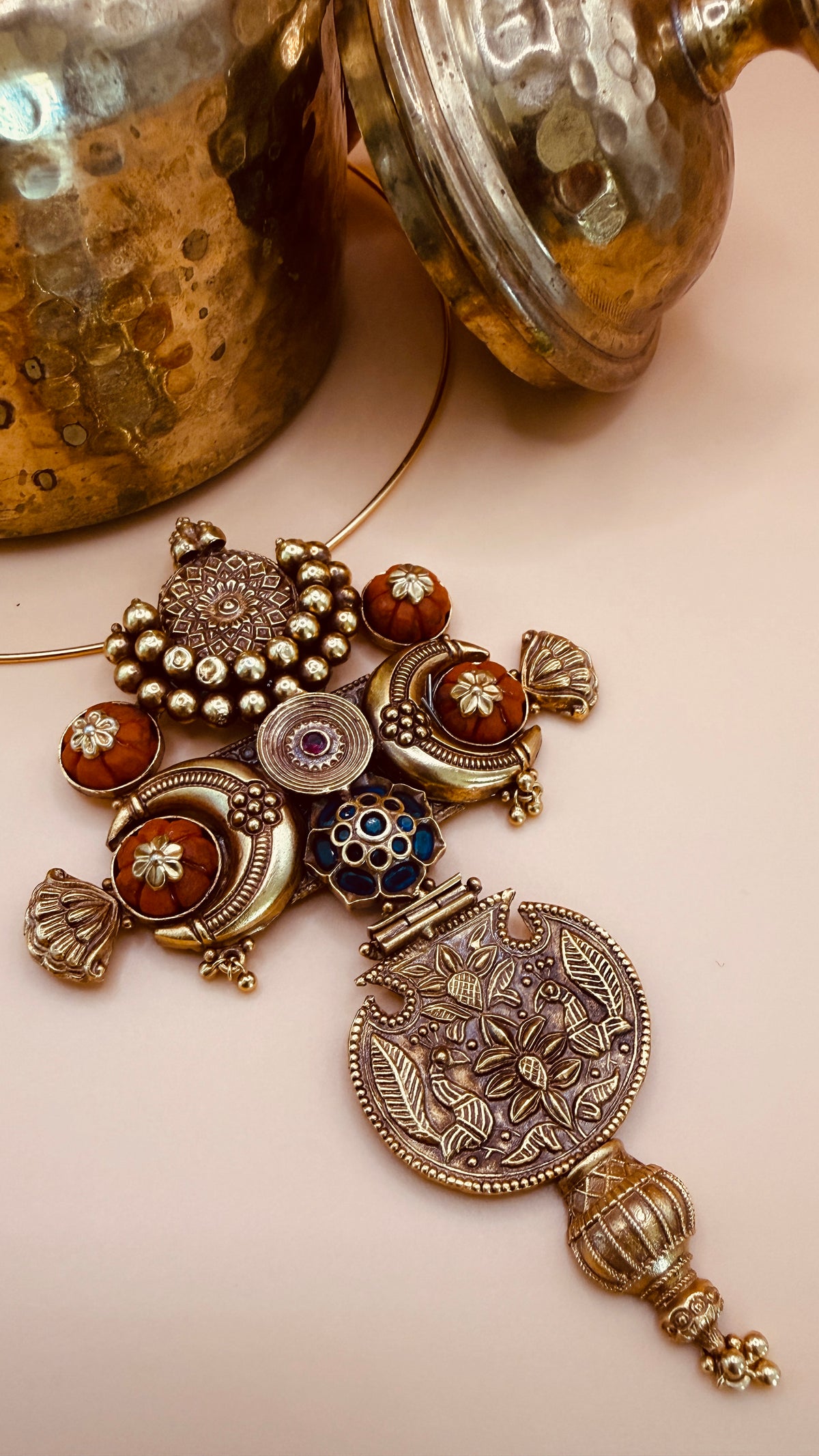 An ornate traditional Indian silver pendant necklace with red, blue, and white enamel details, displayed on a light pink surface with a rustic brass pot in the background.