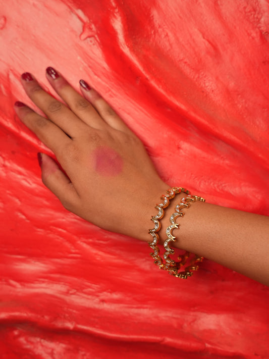 A close-up of a person's hand and wrist wearing three gold crescent moon beaded bracelets, set against a vibrant red textured background.