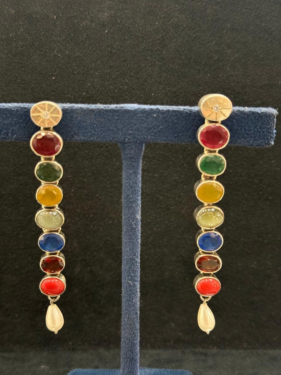A pair of gold chakra dangle earrings with multicolor circular stones and white teardrop pendants, displayed on a blue velvet T-stand against a dark background.