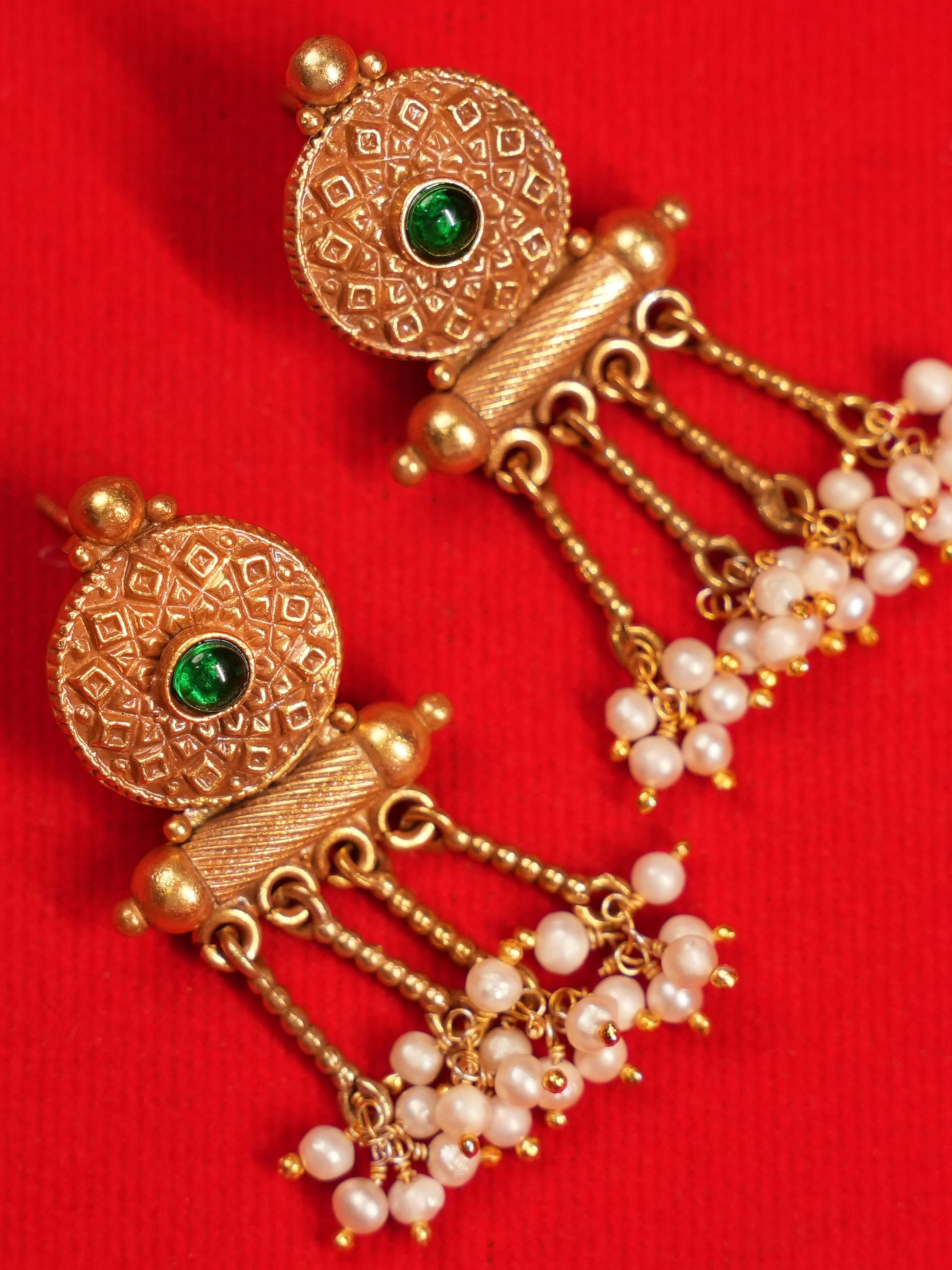 A pair of traditional Indian gold dangle earrings with green stone accents and white pearl beads, displayed on a textured red background.