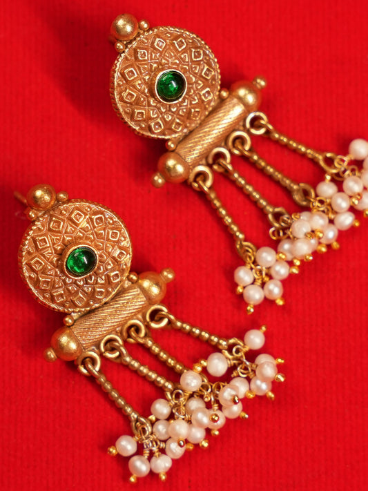 A pair of traditional Indian gold dangle earrings with green stone accents and white pearl beads, displayed on a textured red background.