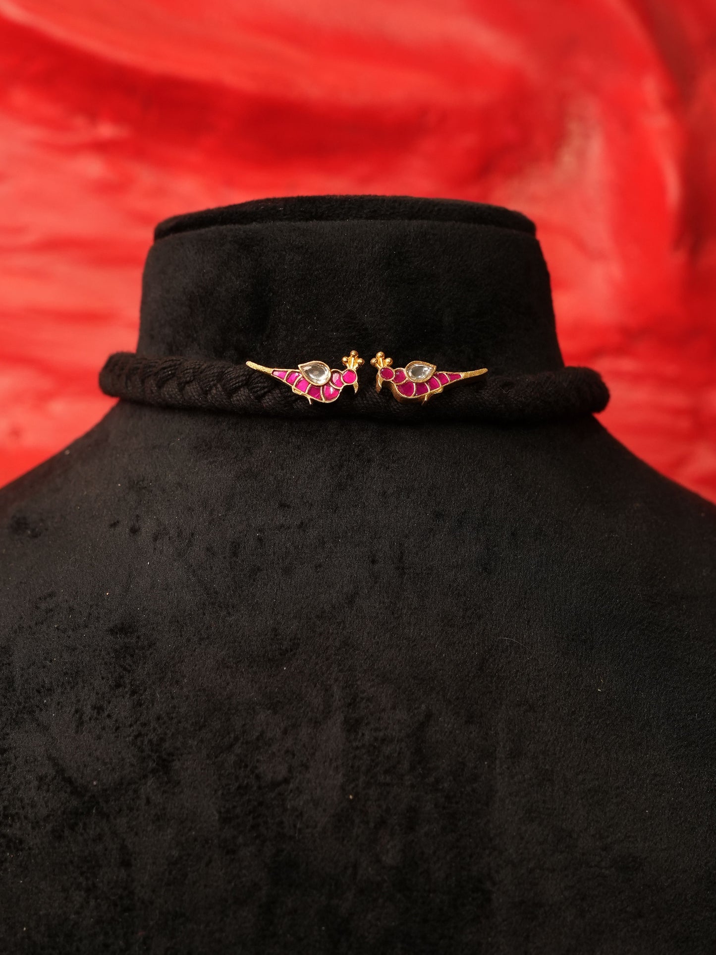 A black braided hair accessory with two gold-toned bird charms embellished with pink and green enamel, displayed on a black velvet mannequin bust against a red fabric background.