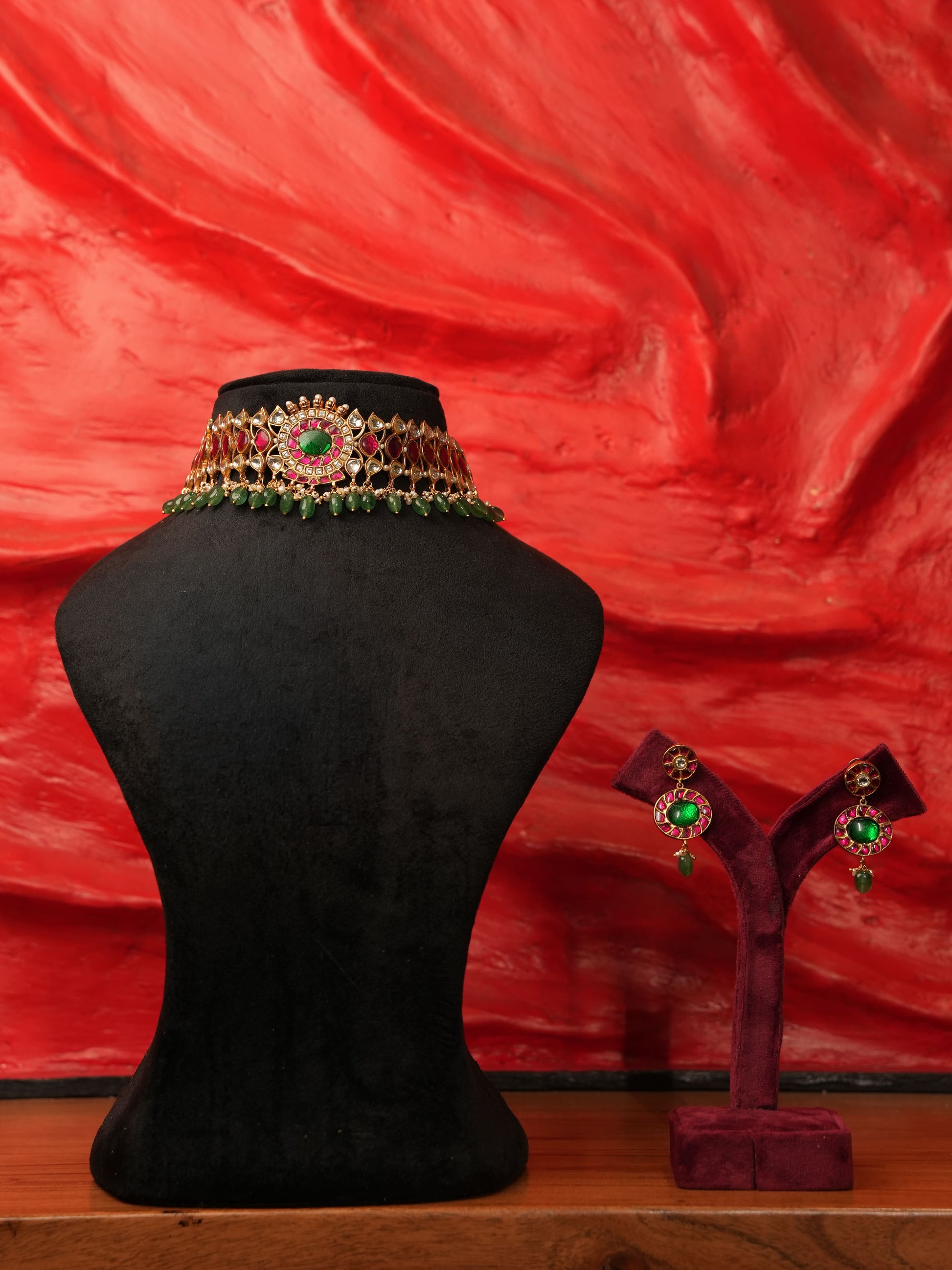 An ornate gold-toned choker necklace with green and red stones is displayed on a black mannequin bust, with matching drop earrings on a small stand against a textured red background.